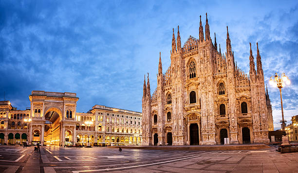 A panoramic view of Milan Cathedral and the adjacent Galleria Vittorio Emanuele II at dusk. The cathedrals Gothic architecture is illuminated under a blue, cloudy sky, with the surrounding area quiet and largely empty.