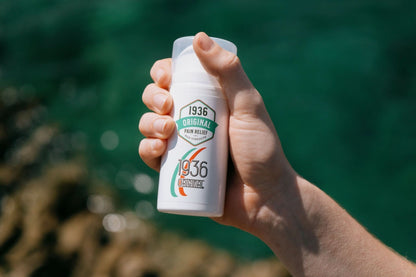 A hand holds a bottle labeled 1936 Original, highlighting the natural formula of the Original Pain Cream against a softly blurred backdrop of tranquil scenery near water. The white bottle, adorned with green and red accents, suggests it’s crafted to effectively soothe back pain, joint discomfort, knee pain, neck issues, and muscle aches.